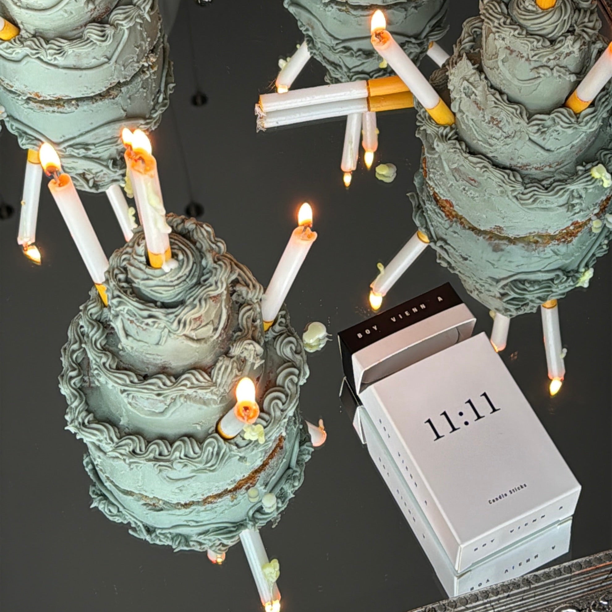 Three tiered cake-shaped candles with lit white and yellow taper candles, arranged on a reflective surface next to a white box marked 11:11.